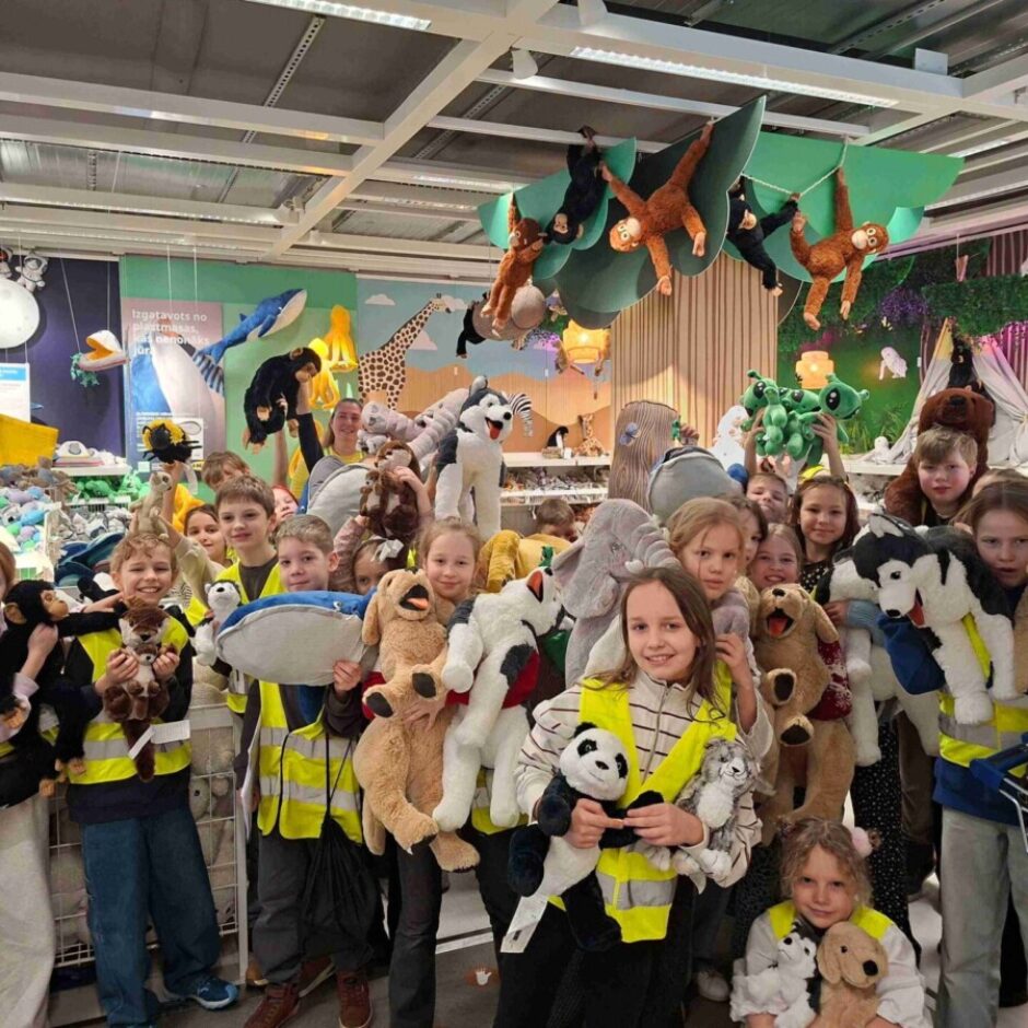 Liela bērnu grupa smaidot pozē IKEA veikalā mīksto rotaļlietu nodaļā. Daudzi bērni valkā dzeltenas vestes un katrs tur rokās vairākas plīša rotaļlietas. Fonā redzami dekoratīvie displeji ar mīkstajiem dzīvniekiem, tostarp pakārtiem pērtiķiem.
