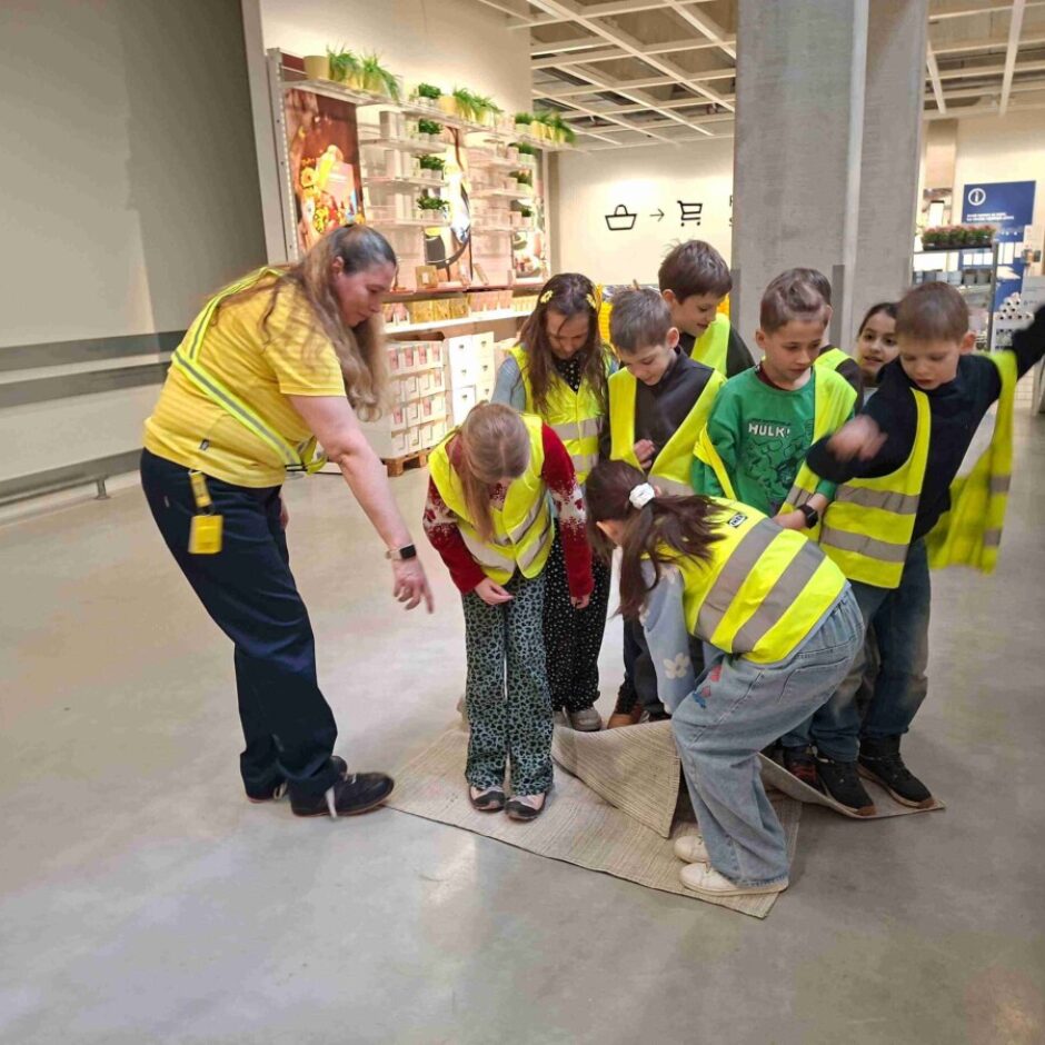 Pieauguša sieviete dzeltenā vestē rāda uz paklāju uz grīdas, ap viņu pulcējušies bērni dzeltenās drošības vestēs skatās uz leju. Viena meitene noliekusies pie paklāja. Viņi atrodas lielā veikalā, fonā redzamas iepirkumu groza un somas ikonas, kā arī informācijas simbols.