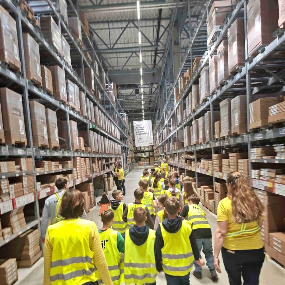 Bērnu un pieaugušo grupa, valkājot dzeltenas atstarojošas vestes, iet cauri plašai noliktavai ar augstiem, kastēm pildītiem plauktiem abās pusēs.