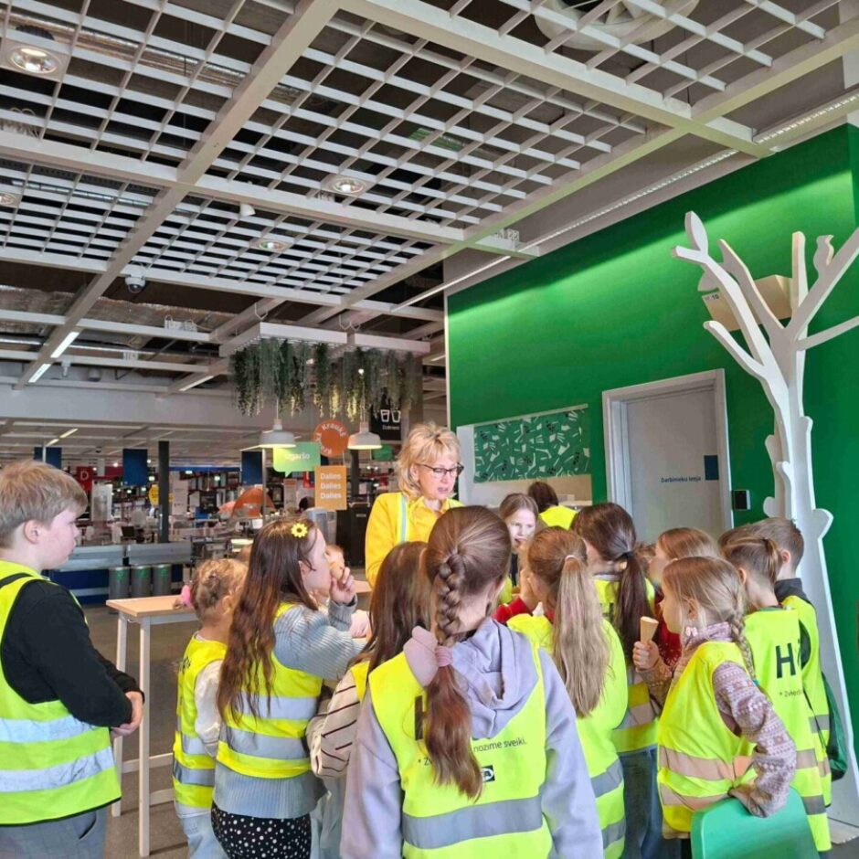 Sieviete dzeltenā vestē sarunājas ar bērnu grupu, kuri arī tērpti dzeltenās atstarojošās vestēs. Viņi atrodas lielā telpā ar zaļu sienu, pie kuras stāv dekoratīvs balts koks.