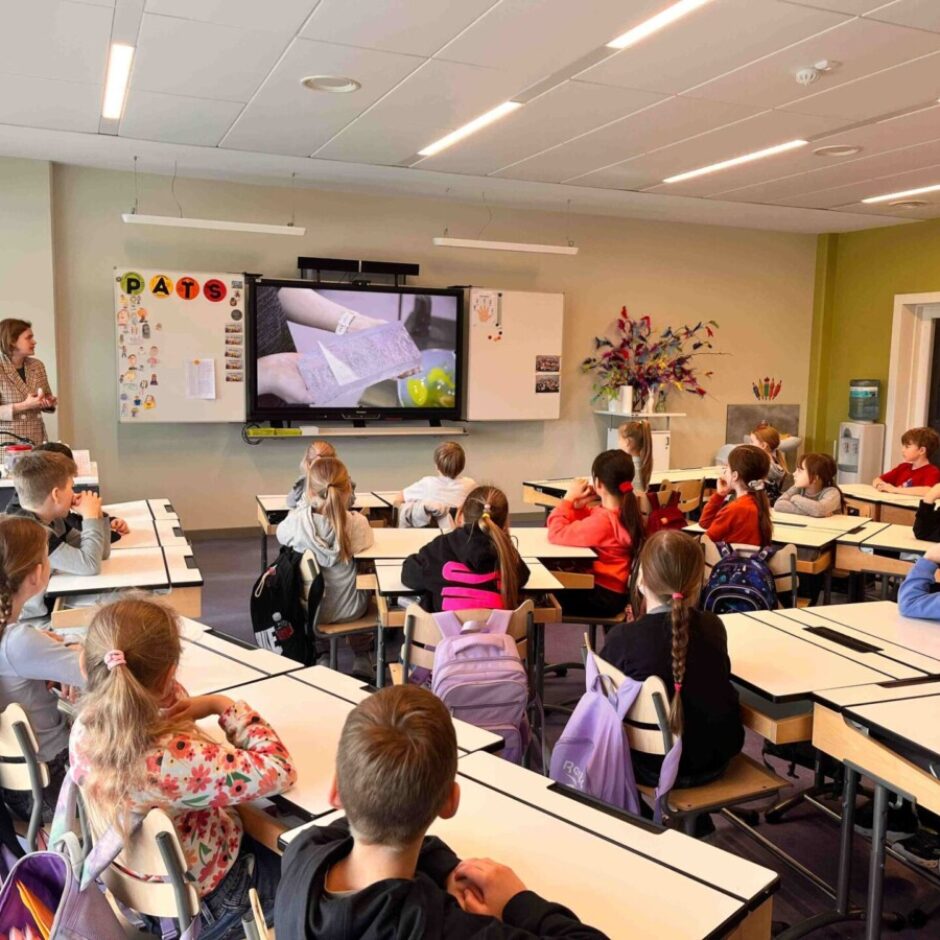 Skolotāja stāv gaišā klasē pie interaktīvā ekrāna, uz kura redzamas rokas, kas veido amatniecības darbu. Vairāki skolēni sēž pie galdiem un uzmanīgi klausās. Uz baltās tāfeles redzami burti 