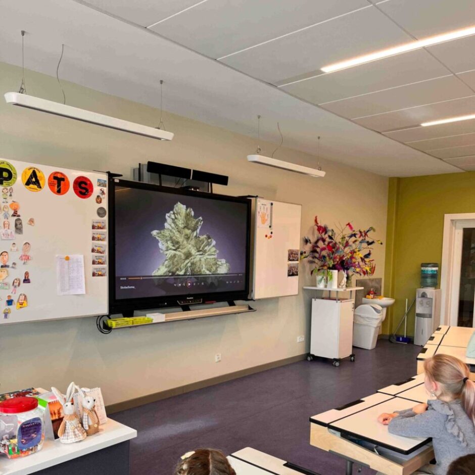 Bērni sēž klasē pie galdiem, skatoties uz interaktīvo tāfeli, kurā redzams palielināts, sarežģīts zaļš veidojums, iespējams, augs vai minerāls.