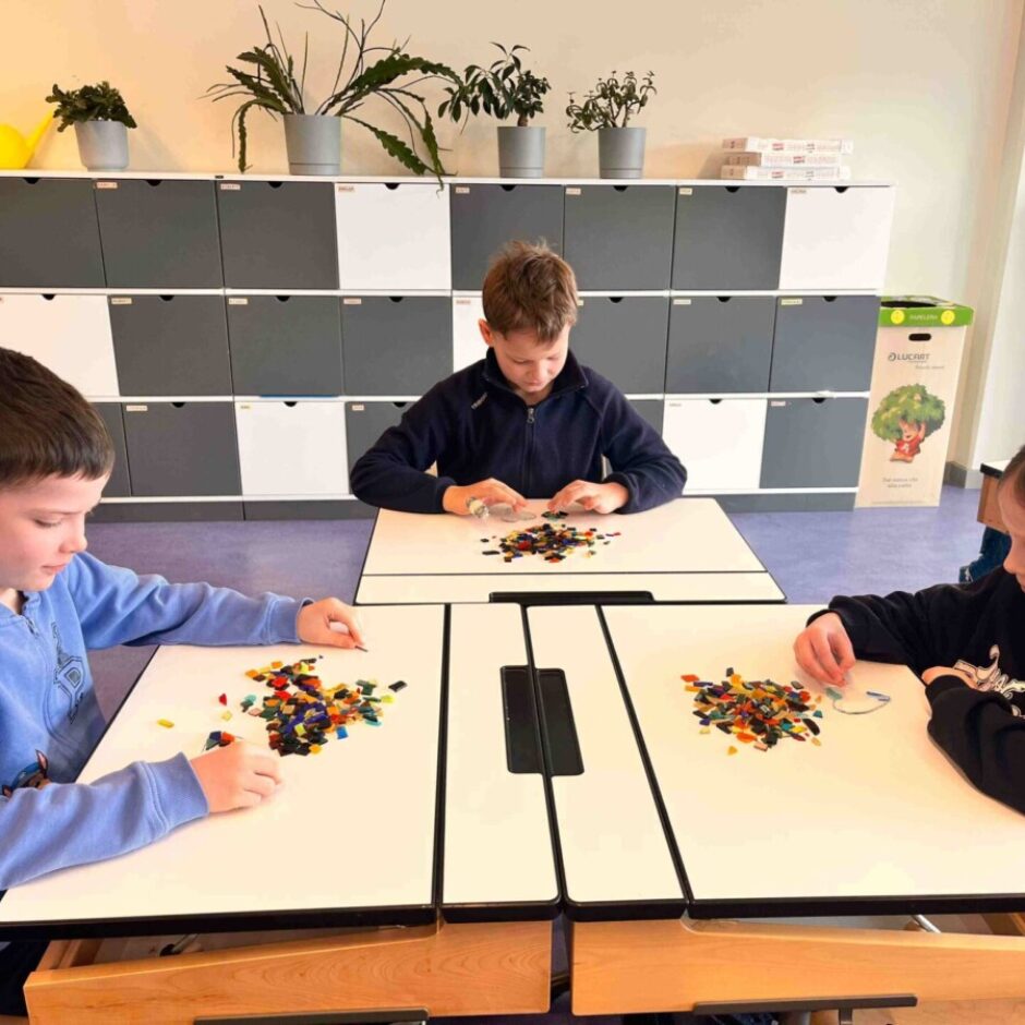 Trīs bērni (divi zēni un meitene) klasē pie galdiem spēlējas ar krāsainiem stikliņiem