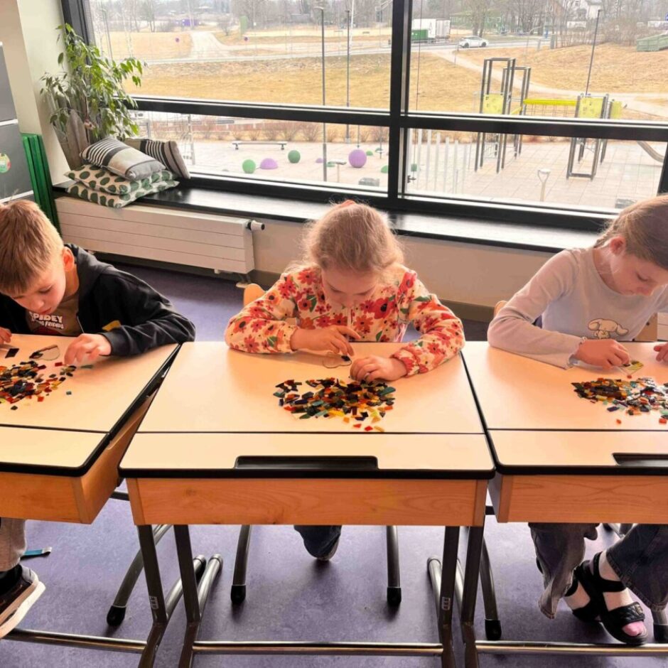 Trīs bērni, divas meitenes un zēns, sēž pie galdiem un veido mozaīkas no maziem krāsainiem gabaliņiem