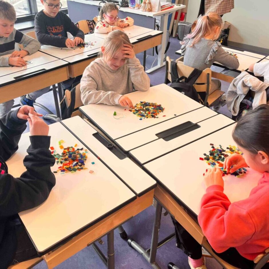 Vairāki bērni sēž pie klases galdiem un cītīgi strādā ar mazām, krāsainām caurspīdīgām amatniecības detaļām.