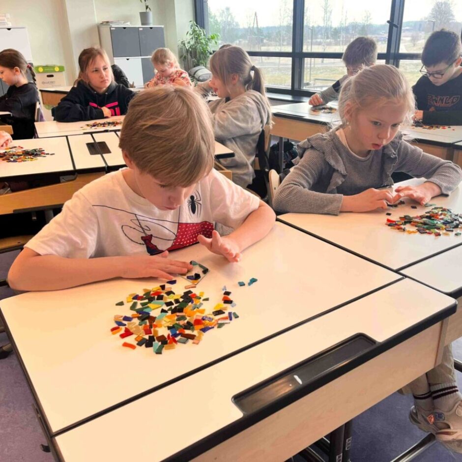 Vairāki bērni sēž klasē pie galdiem un darbojas ar krāsainiem mozaīkas gabaliņiem. Divi bērni priekšplānā uzmanīgi kārto fragmentus uz saviem galdiem.