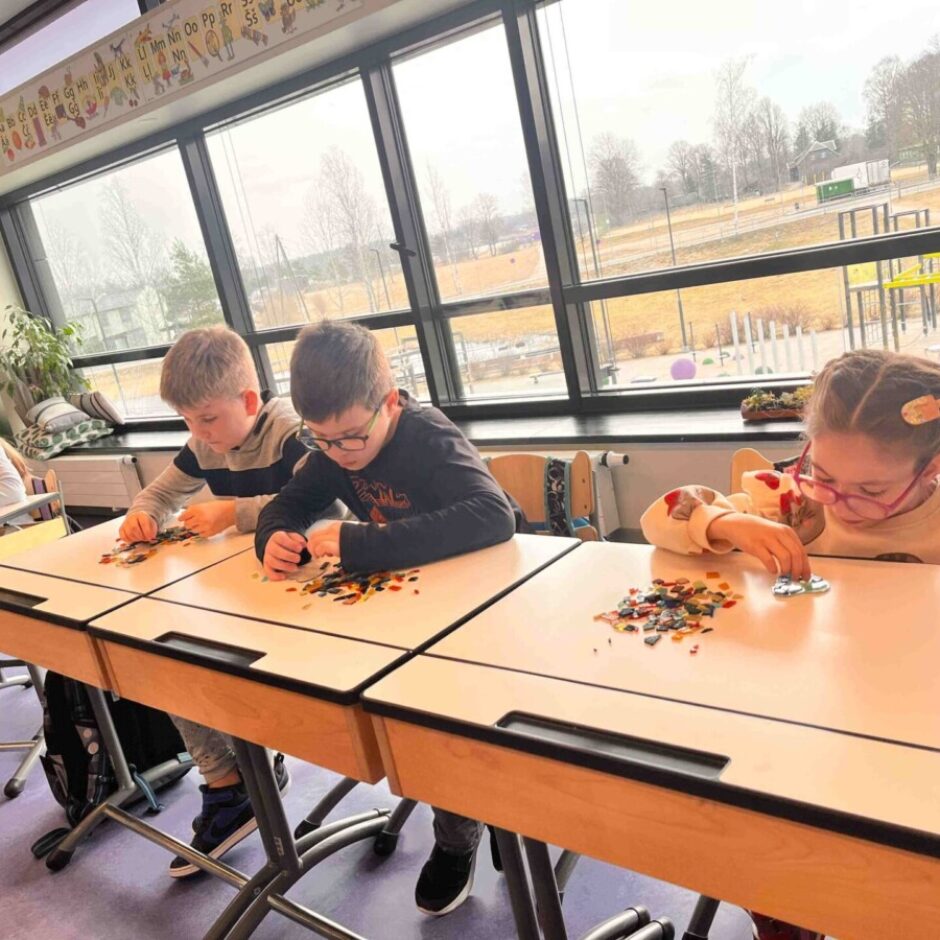 Četri bērni klasē sēž pie galdiem, koncentrējoties uz mazu, krāsainu gabaliņu šķirošanu vai kārtošanu uz savām virsmām. Aiz viņiem ir lieli logi ar skatu uz ārpusi ar kokiem un rotaļu laukumu. Virs logiem ir redzams alfabēta plakāts