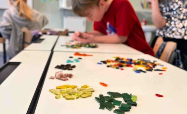 Bērni radošā nodarbībā veido mozaīku. Zēns sarkanā kreklā koncentrējas uz darbu, izvietojot krāsainus stikla gabaliņus uz balta galda. Priekšplānā redzamas sakārtotas kaudzītes ar zilas, zaļas, dzeltenas un brūnas krāsas stikla gabaliņiem.