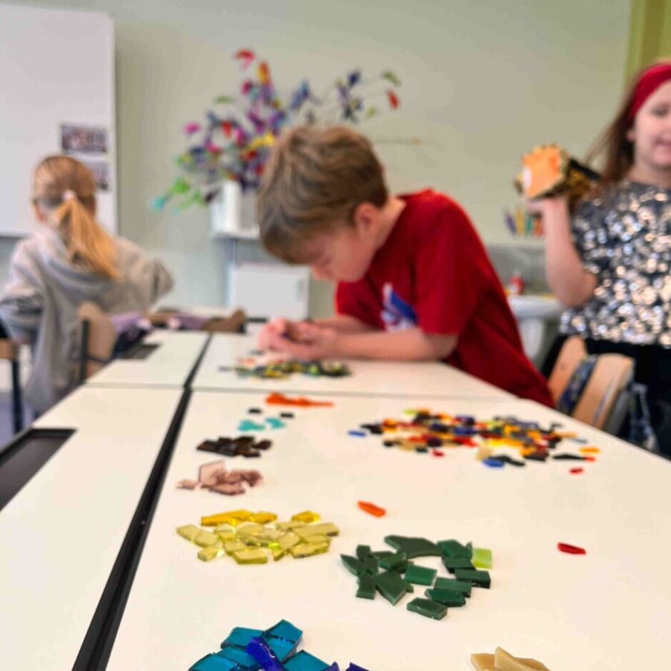 Bērni radošā nodarbībā veido mozaīku. Zēns sarkanā kreklā koncentrējas uz darbu, izvietojot krāsainus stikla gabaliņus uz balta galda. Priekšplānā redzamas sakārtotas kaudzītes ar zilas, zaļas, dzeltenas un brūnas krāsas stikla gabaliņiem.