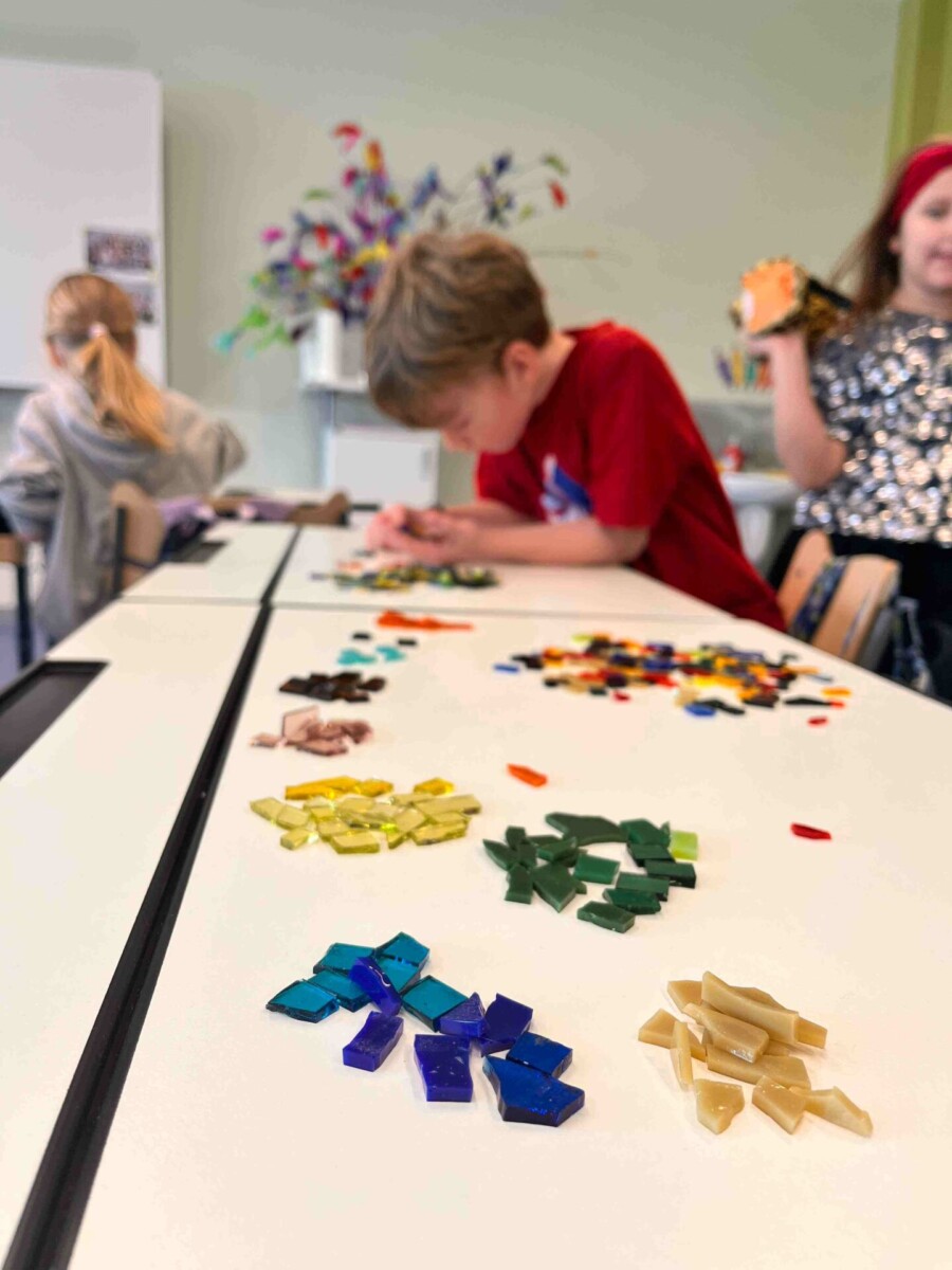 Bērni radošā nodarbībā veido mozaīku. Zēns sarkanā kreklā koncentrējas uz darbu, izvietojot krāsainus stikla gabaliņus uz balta galda. Priekšplānā redzamas sakārtotas kaudzītes ar zilas, zaļas, dzeltenas un brūnas krāsas stikla gabaliņiem.