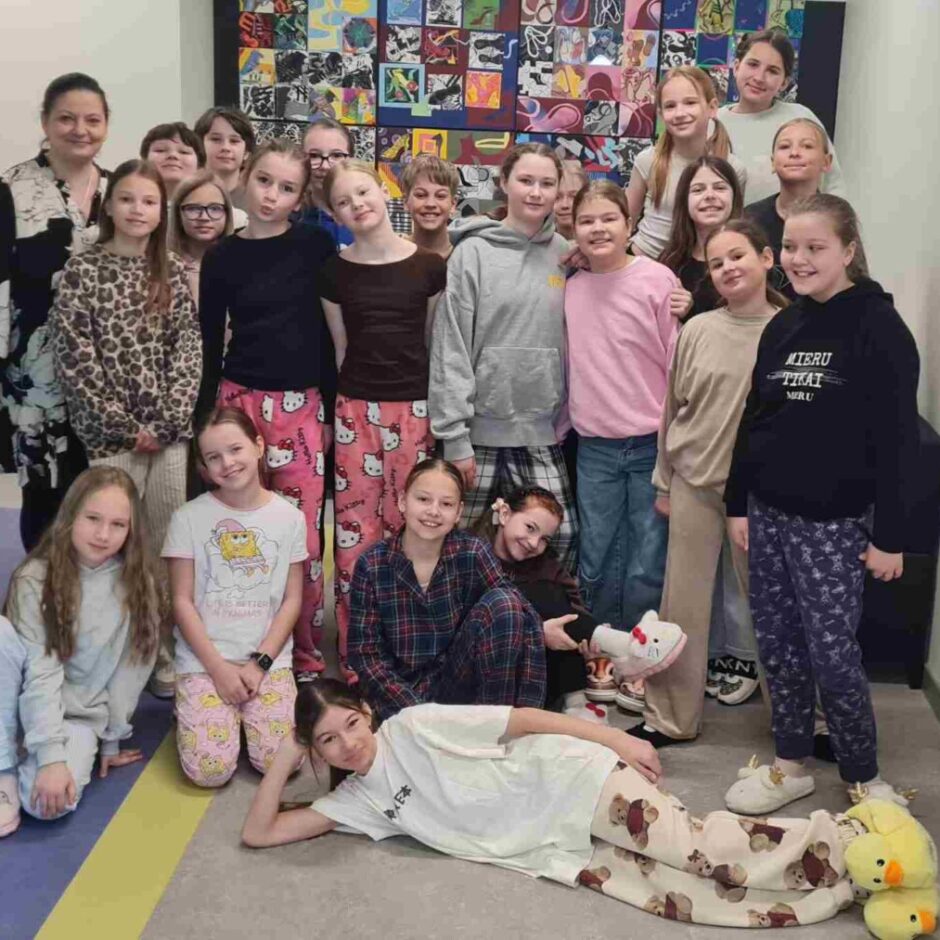 Liela meiteņu grupa un viena pieaugusi sieviete smaidot pozē iekštelpās, valkājot dažādas pidžamas un čības.