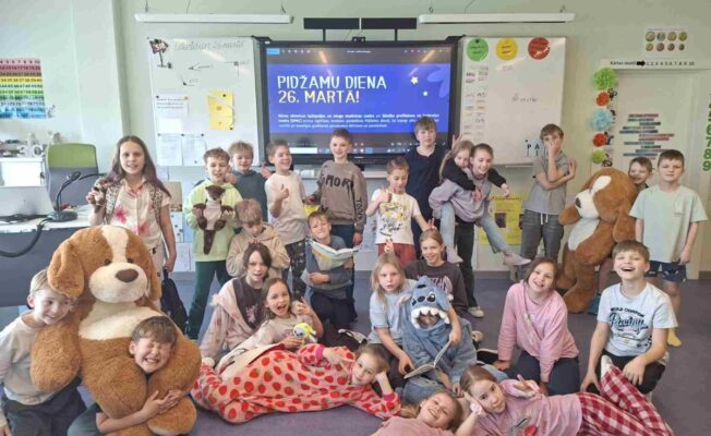 smaidoši bērni pozē skolas klasē, daudzi no viņiem ir ģērbušies pidžamās un tur rokās mīkstās rotaļlietas. Fonā uz interaktīvā ekrāna redzams teksts: 