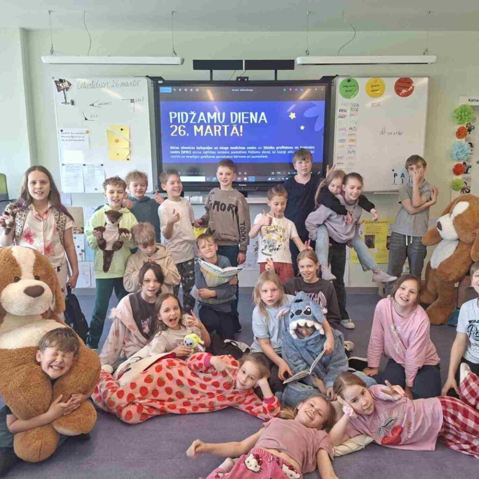 smaidoši bērni pozē skolas klasē, daudzi no viņiem ir ģērbušies pidžamās un tur rokās mīkstās rotaļlietas. Fonā uz interaktīvā ekrāna redzams teksts: 