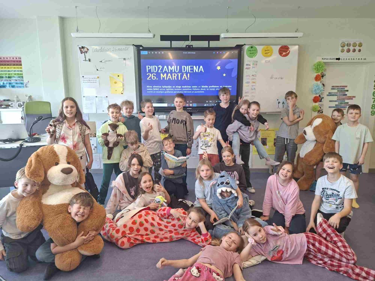 smaidoši bērni pozē skolas klasē, daudzi no viņiem ir ģērbušies pidžamās un tur rokās mīkstās rotaļlietas. Fonā uz interaktīvā ekrāna redzams teksts: 