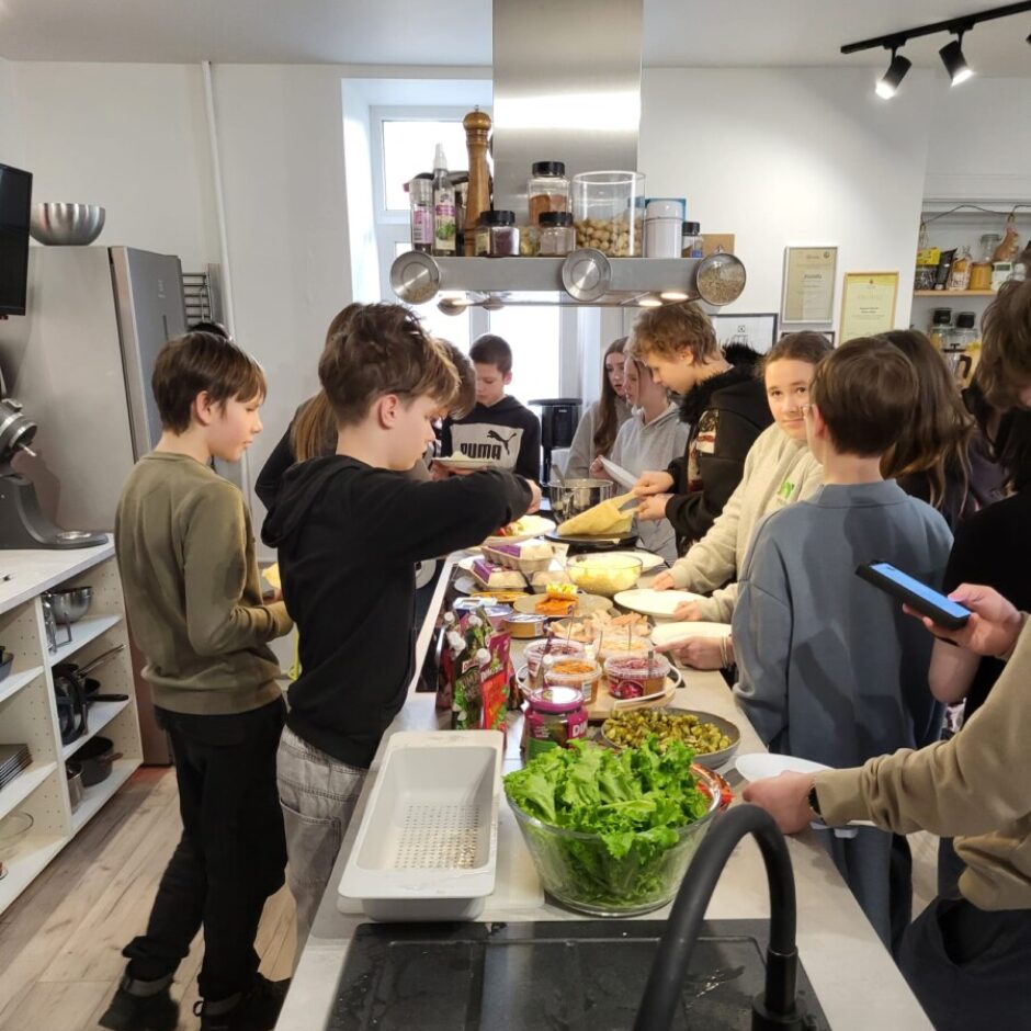 Grupa bērnu un pusaudžu modernā virtuvē apkalpo sevi no bufetes galda, liekot ēdienu uz šķīvjiem. Uz galda ir dažādi ēdieni, tostarp liela bļoda ar salātiem. Priekšplānā redzama melna izlietne.