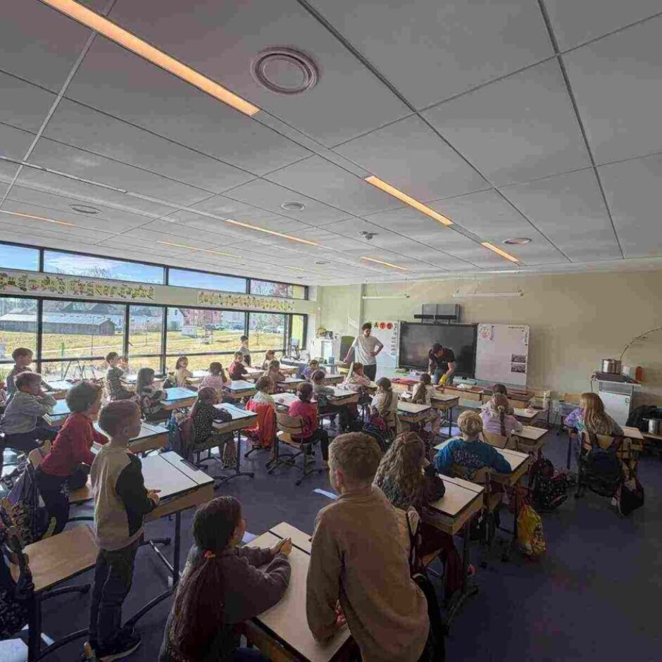 Mūsdienīga skolas klase ar lieliem logiem, caur kuriem redzami lauki un ēkas tālumā. Klasē daudzi bērni sēž pie galdiem, bet daži stāv vai pārvietojas. Pie klases priekšējās sienas divi pieaugušie aktīvi nodarbojas ar bērniem blakus tāfelei. Virs logiem ir krāsains dekoratīvs baneris ar burtiem.