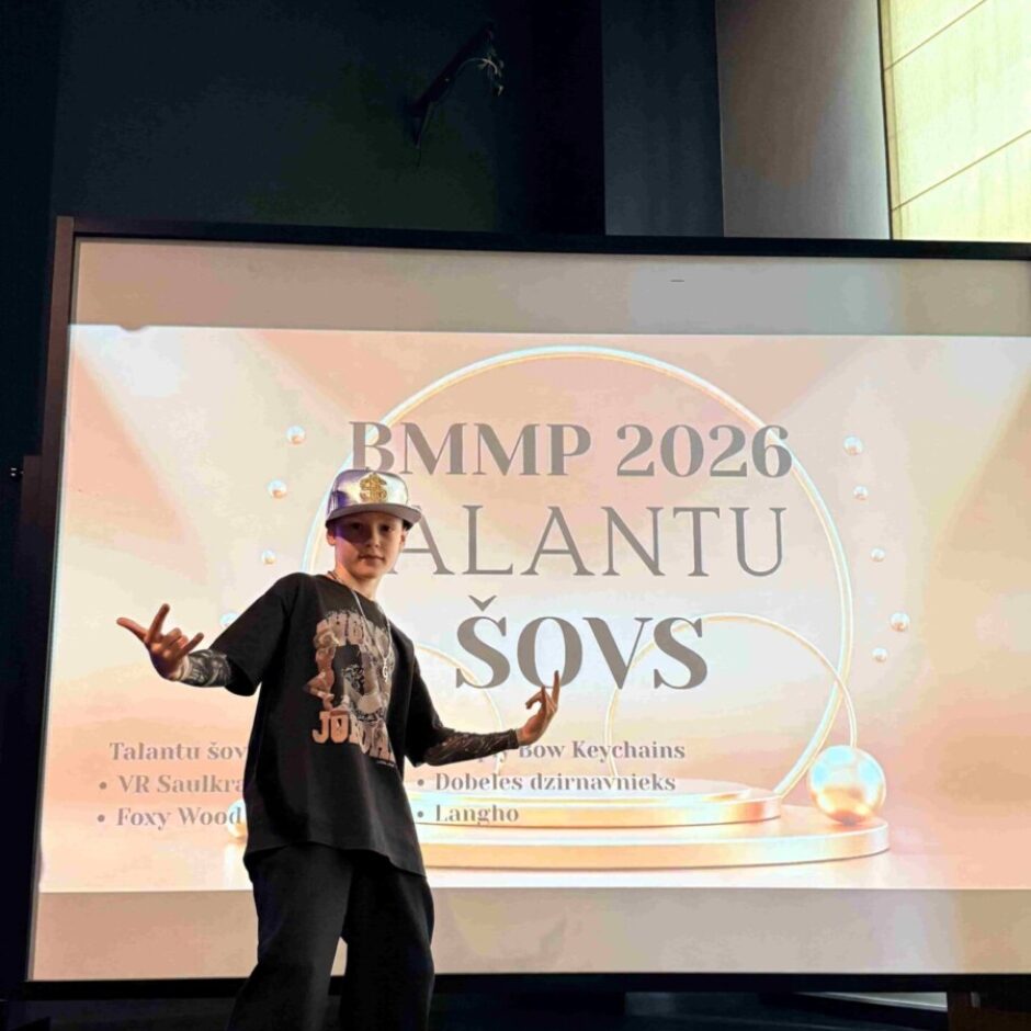 Jauns zēns uz skatuves pozē ar paceltām rokām un cepuri ar nagu uz aizmuguri, melnā T-kreklā un platās biksēs. Aiz viņa redzams liels ekrāns ar uzrakstu 