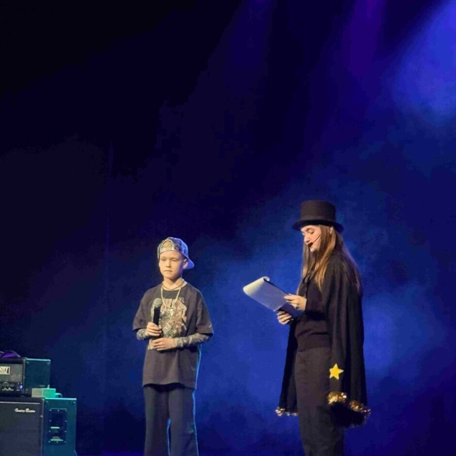 Uz skatuves ar zilu apgaismojumu jauns zēns ar beisbola cepuri tur mikrofonu, blakus meitene melnā apmetnī ar zelta zvaigzni un cilindra cepurē tur baltu planšeti. Viņi, iespējams, uzstājas.