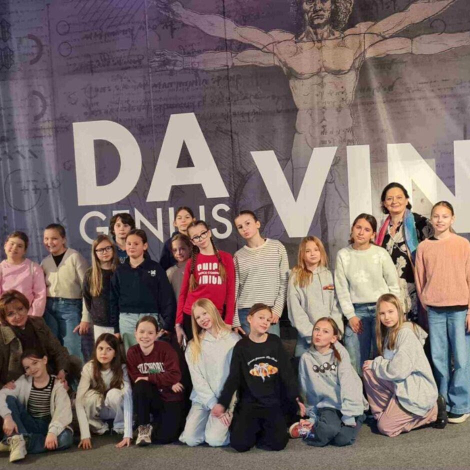 Grupa bērnu un divas sievietes pozē lielas sienas priekšā ar uzrakstu 