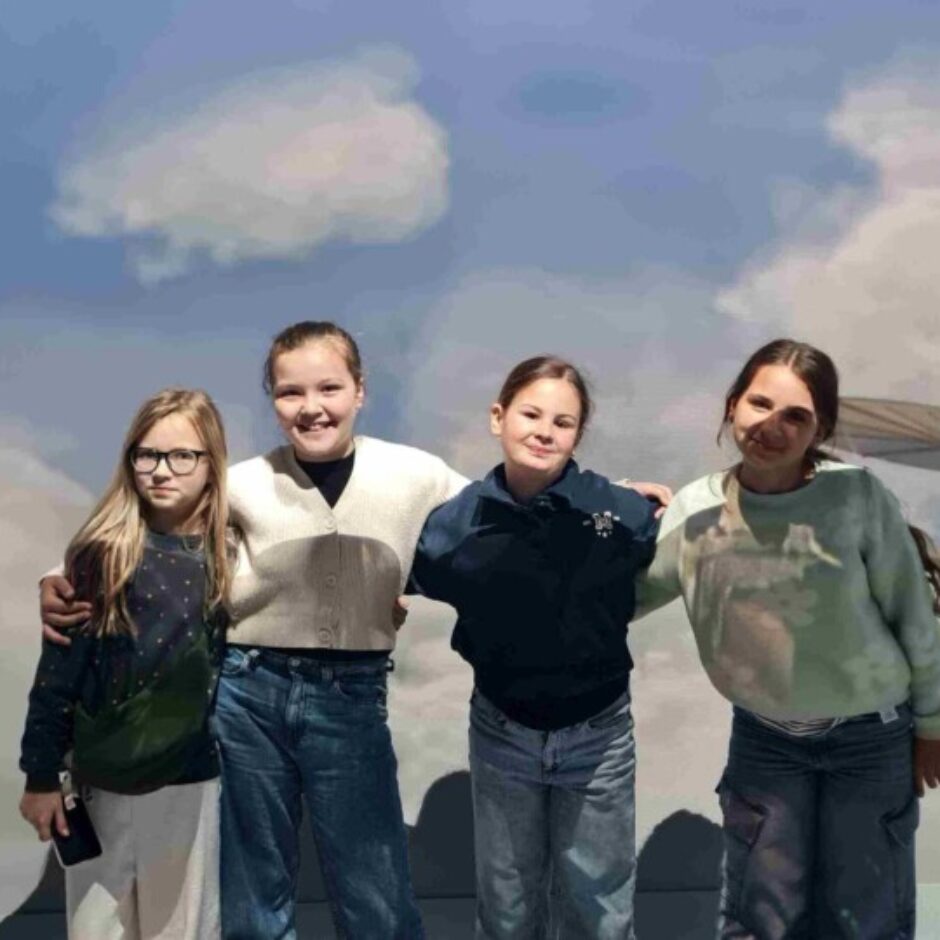 Četras jaunas, smaidīgas meitenes, apskāvušās, pozē pie fona ar mākoņainām debesīm un diviem stilizētiem agrīniem lidošanas aparātiem.