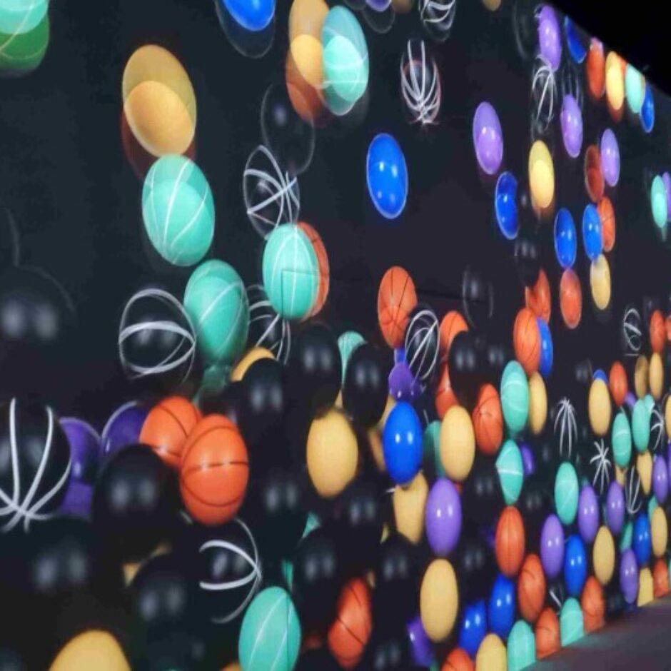 Imersīva telpa, kuras sienās projicētas daudzas krāsainas bumbas, tostarp basketbola bumbas un vienkrāsainas bumbas zilā, oranžā, melnā, tirkīza, dzeltenā un violetā krāsā, kas dinamiski pārvietojas pa tumšu fonu. Labajā pusē redzams cilvēka siluets.