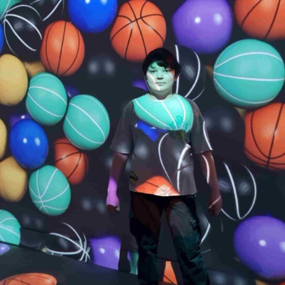 Jauns cilvēks stāv tumšā telpā, ko apgaismo daudzu krāsainu basketbola bumbu un ovālu formu projekcijas uz sienām un grīdas. Krāsainās projekcijas redzamas arī uz cilvēka sejas un krekla.