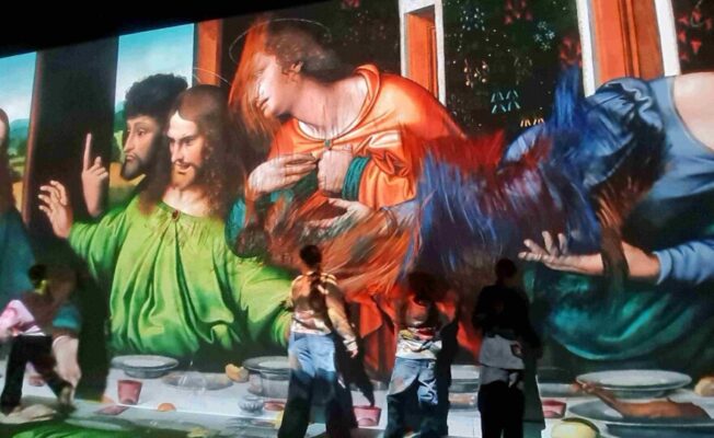 Ieskaujoša projekcija attēlo lielformāta gleznu ar Bībeles ainu, piemēram, Pēdējās vakariņas, uz sienas un grīdas. Redzami daudzi tēli pie galda, un vairāki cilvēki stāv priekšā projekcijai, vērojot to.