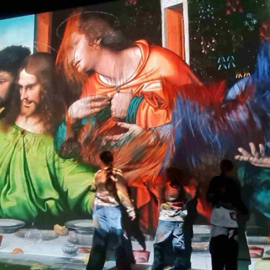 Ieskaujoša projekcija attēlo lielformāta gleznu ar Bībeles ainu, piemēram, Pēdējās vakariņas, uz sienas un grīdas. Redzami daudzi tēli pie galda, un vairāki cilvēki stāv priekšā projekcijai, vērojot to.