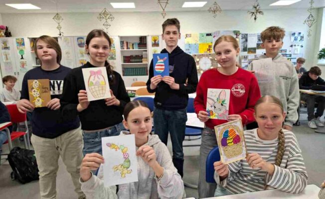 Septiņi skolēni klasē, četri no tiem ir meitenes un trīs zēni, turot rokās dažādus paštaisītus Lieldienu apsveikumus un zīmējumus. Attēlos redzami Lieldienu zaķi, krāsainas olas grozos un viens zīmējums ar olām, kas izskatās pēc cepumiem. Uz vienas kartītes ir uzraksts 
