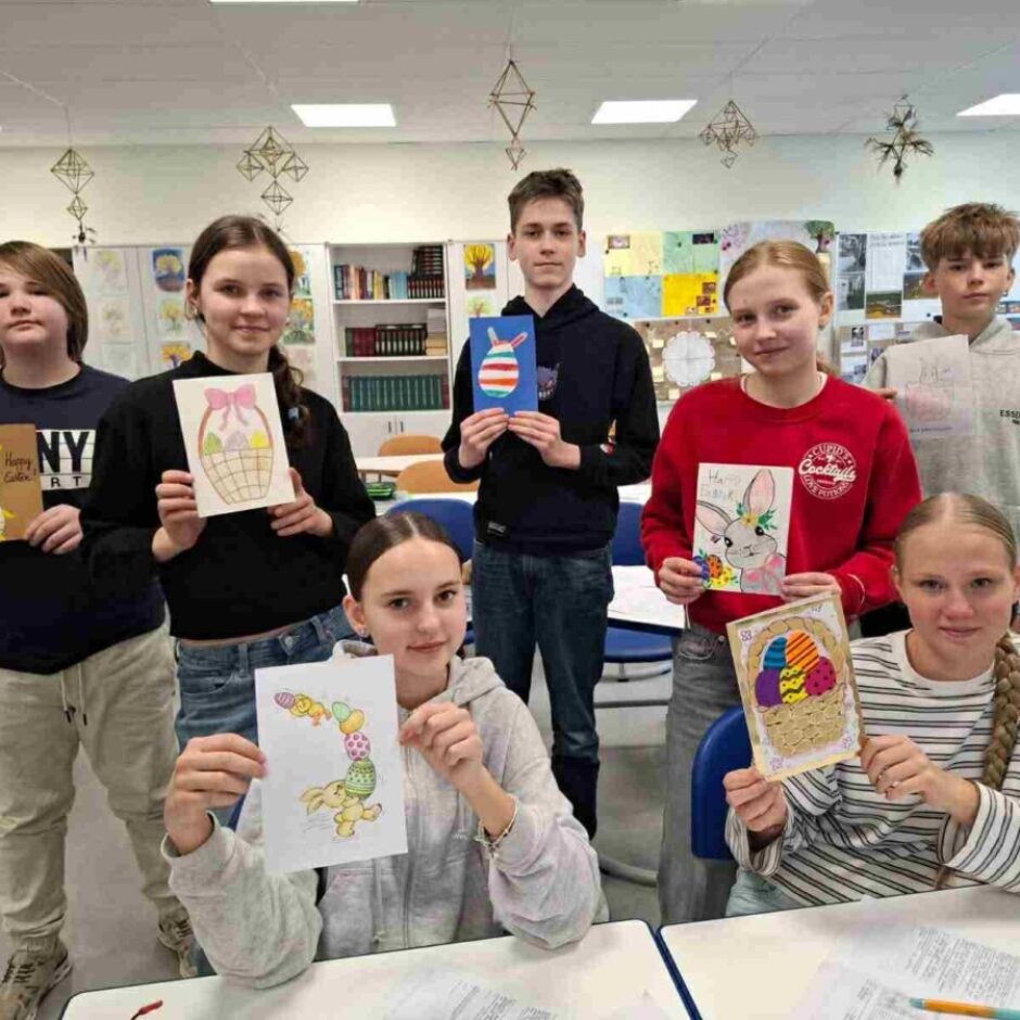 Septiņi skolēni klasē, četri no tiem ir meitenes un trīs zēni, turot rokās dažādus paštaisītus Lieldienu apsveikumus un zīmējumus. Attēlos redzami Lieldienu zaķi, krāsainas olas grozos un viens zīmējums ar olām, kas izskatās pēc cepumiem. Uz vienas kartītes ir uzraksts 