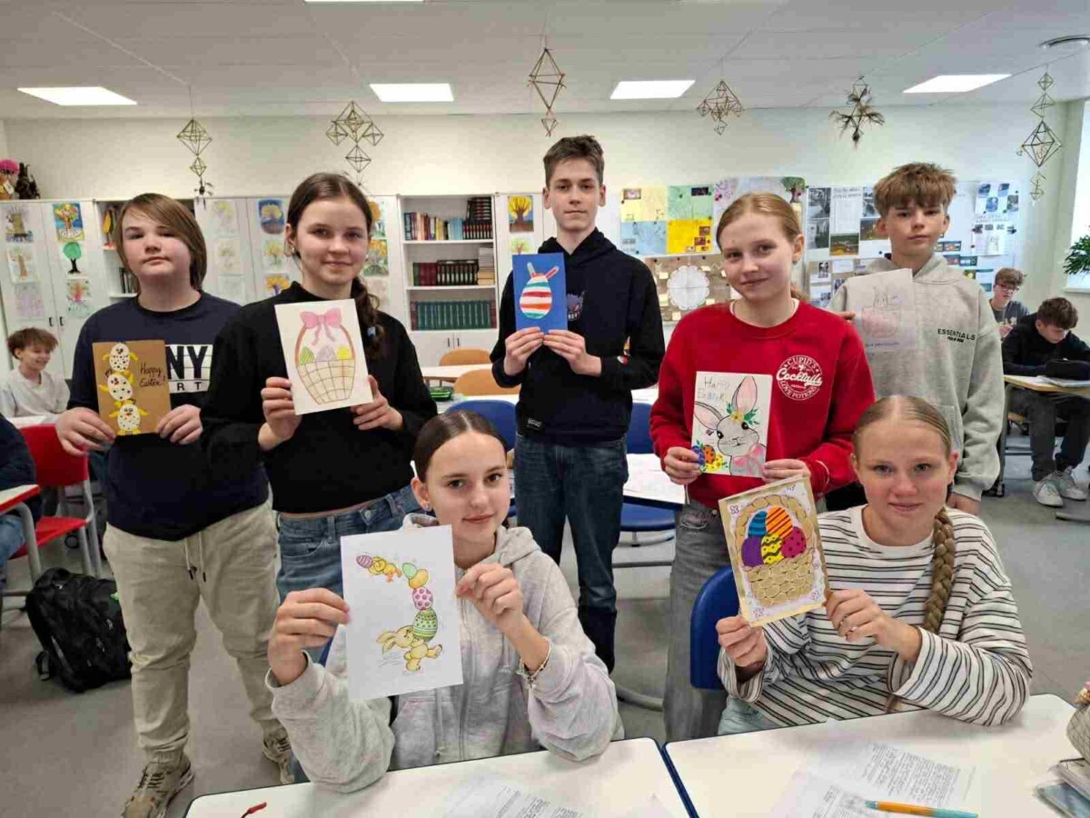 Septiņi skolēni klasē, četri no tiem ir meitenes un trīs zēni, turot rokās dažādus paštaisītus Lieldienu apsveikumus un zīmējumus. Attēlos redzami Lieldienu zaķi, krāsainas olas grozos un viens zīmējums ar olām, kas izskatās pēc cepumiem. Uz vienas kartītes ir uzraksts 