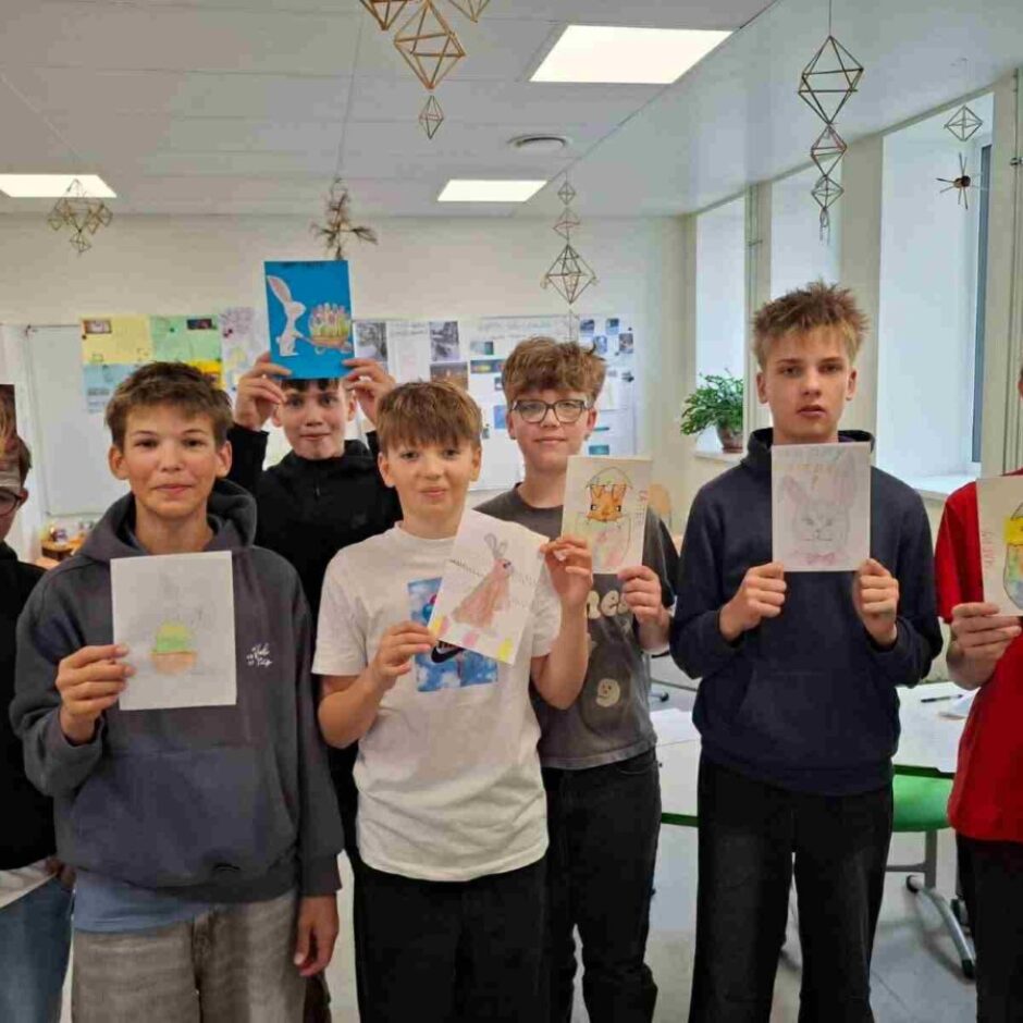 Septiņi zēni klasē tur rokās Lieldienu tematikas zīmējumus, kuros redzami zaķi un olas.