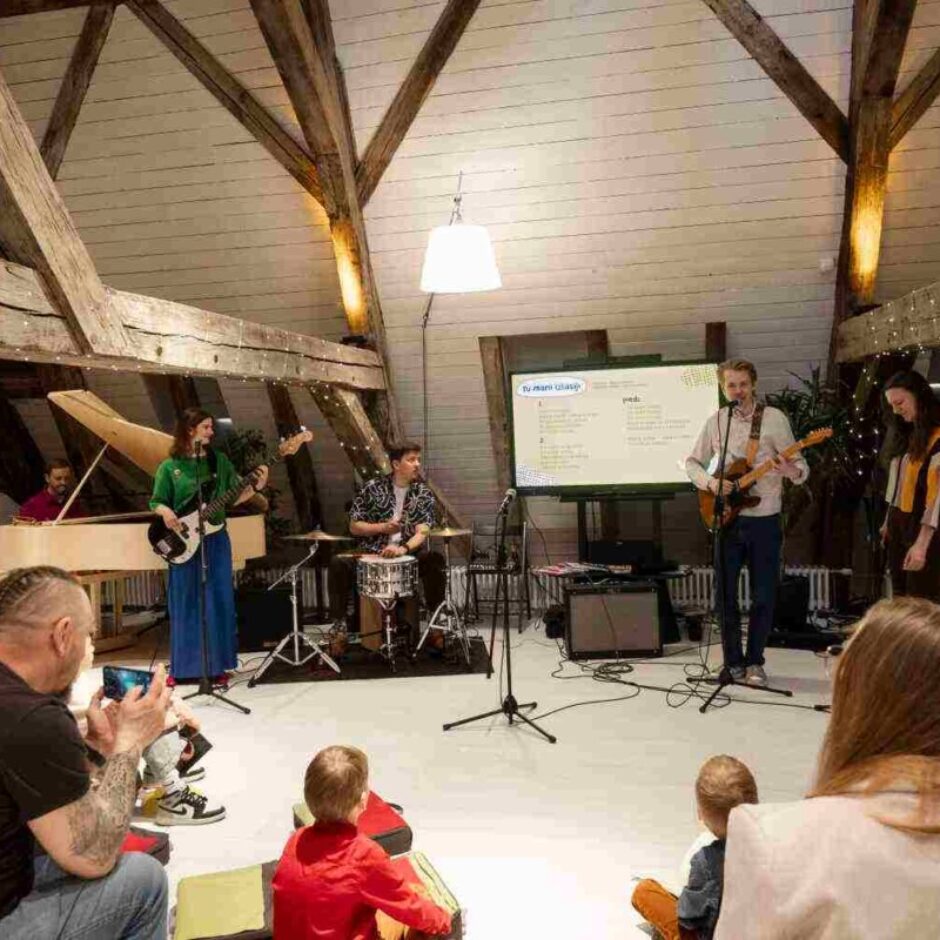 Mūzikas grupa uzstājas bēniņu telpā ar koka sijām, kas izrotātas ar lampiņām. Sieviete spēlē basģitāru, vīrietis bungas un cits vīrietis akustisko ģitāru, dziedot mikrofonā. Aiz grupas ir ekrāns ar uzrakstu 