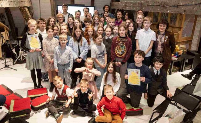 Liela grupa bērnu un daži pieaugušie pozē fotogrāfijai iekštelpās ar koka sijām. Vairāki bērni lepni tur diplomus, uz viena no tiem skaidri redzams vārds 'DIPLOMS', uz cita 'Girls' Day'. Viens bērns valkā džemperi ar uzrakstu 'HOGWARTS'. Atmosfēra ir priecīga un svinīga.