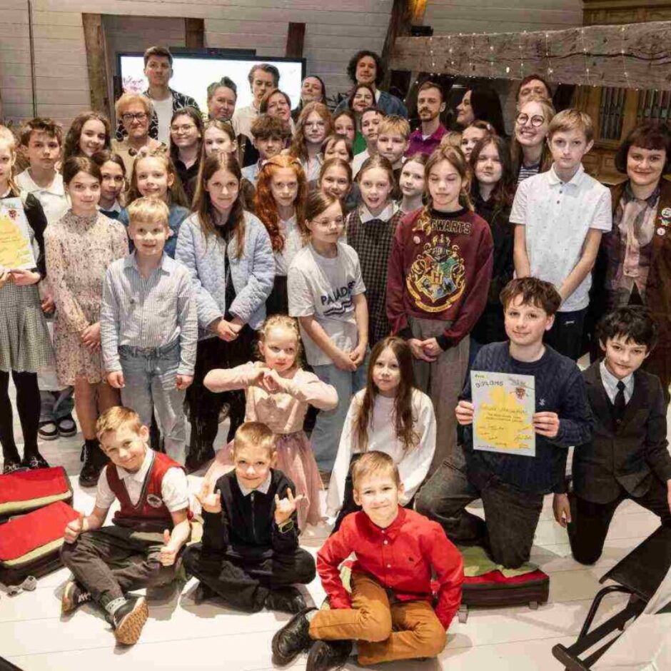 Liela grupa bērnu un daži pieaugušie pozē fotogrāfijai iekštelpās ar koka sijām. Vairāki bērni lepni tur diplomus, uz viena no tiem skaidri redzams vārds 'DIPLOMS', uz cita 'Girls' Day'. Viens bērns valkā džemperi ar uzrakstu 'HOGWARTS'. Atmosfēra ir priecīga un svinīga.