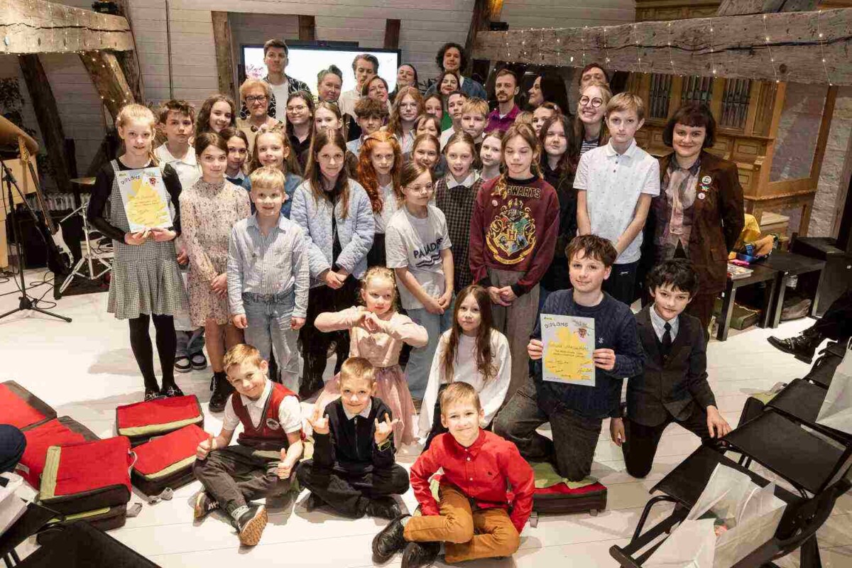 Liela grupa bērnu un daži pieaugušie pozē fotogrāfijai iekštelpās ar koka sijām. Vairāki bērni lepni tur diplomus, uz viena no tiem skaidri redzams vārds 'DIPLOMS', uz cita 'Girls' Day'. Viens bērns valkā džemperi ar uzrakstu 'HOGWARTS'. Atmosfēra ir priecīga un svinīga.