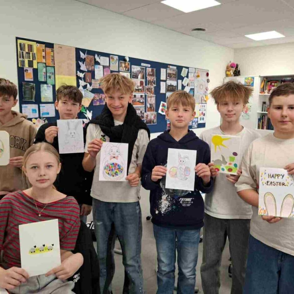 Grupa bērnu, septiņi zēni un viena meitene, stāv klasē un smaida, turot rokās pašu zīmētus Lieldienu attēlus vai apsveikuma kartītes ar zaķiem, olām un cāļiem. Uz vienas kartītes ir uzraksts 