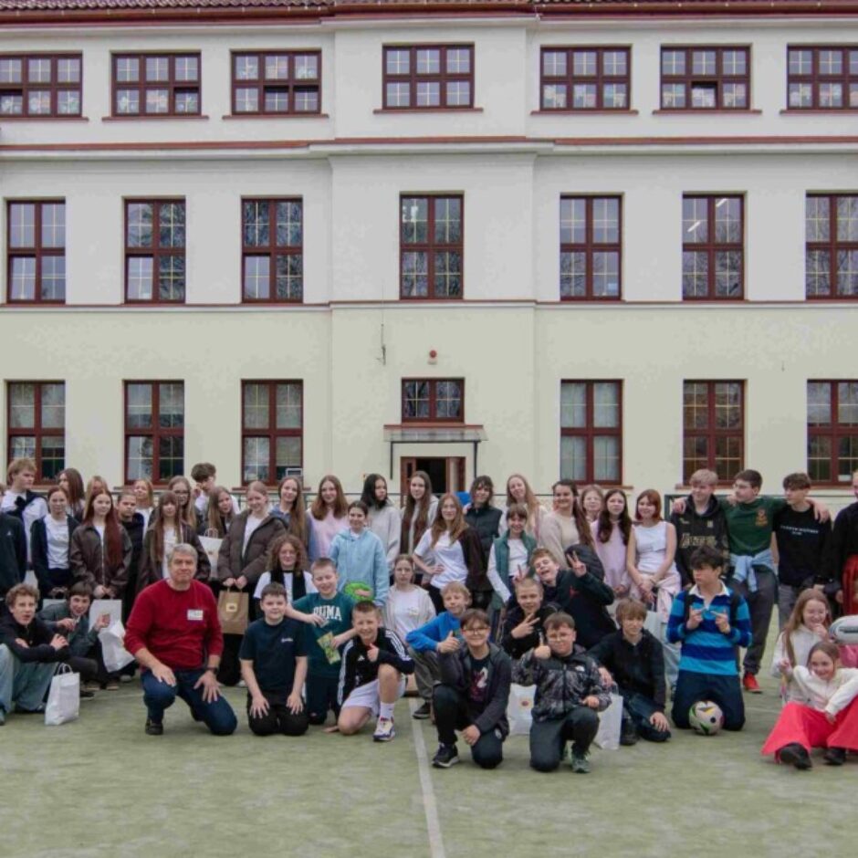Liela skolēnu un pieaugušo grupa pozē kopīgai fotogrāfijai gaišas skolas ēkas priekšā uz zaļa laukuma. Daudzi smaida, un daži tur rokās somas.