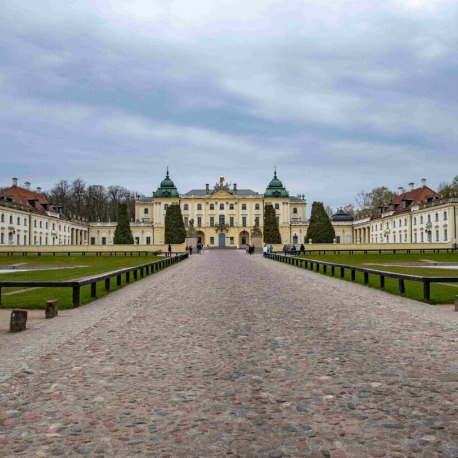 Liela, gaiši dzeltena pils ar simetriskiem spārniem un zaļiem kupoliem, uz kuru ved bruģēts ceļš ar zaļiem mauriņiem abās pusēs. Vairāki cilvēki staigā pa ceļu zem pelēkām, mākoņainām debesīm.