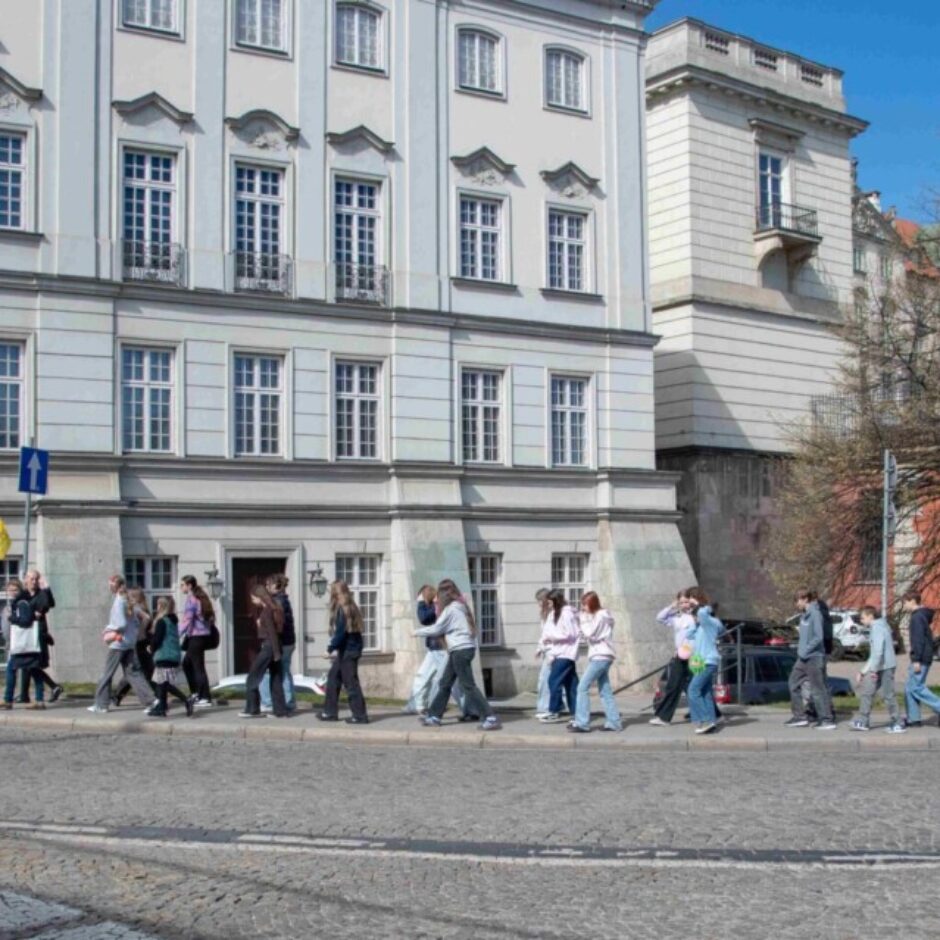 Cilvēku grupa, šķērsojot bruģētu ielu, soļo pa gājēju pāreju gaišas, daudzlogainas vēsturiskas ēkas priekšā. Tālāk redzams kails koks un sarkanu ķieģeļu celtne.