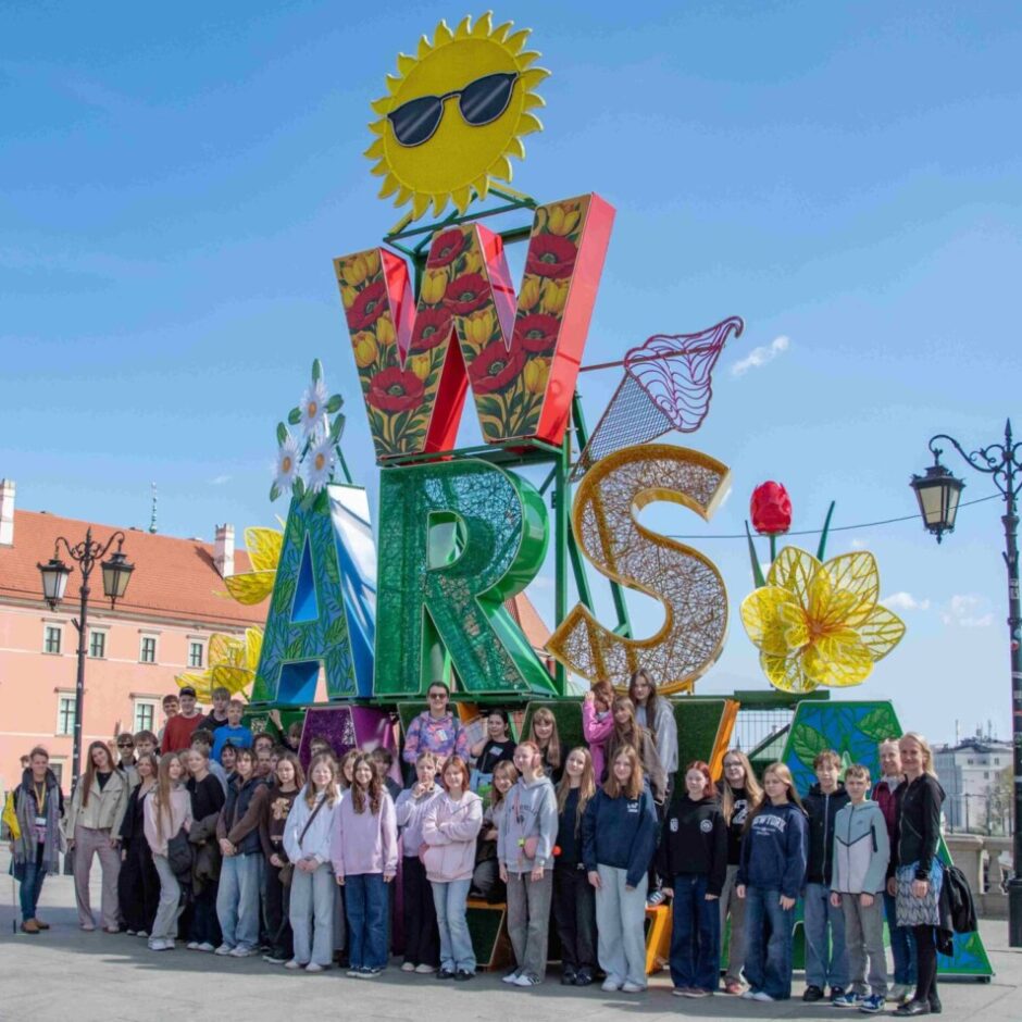 Liela skolēnu un pieaugušo grupa pozē pie milzīga, krāsaina uzraksta 