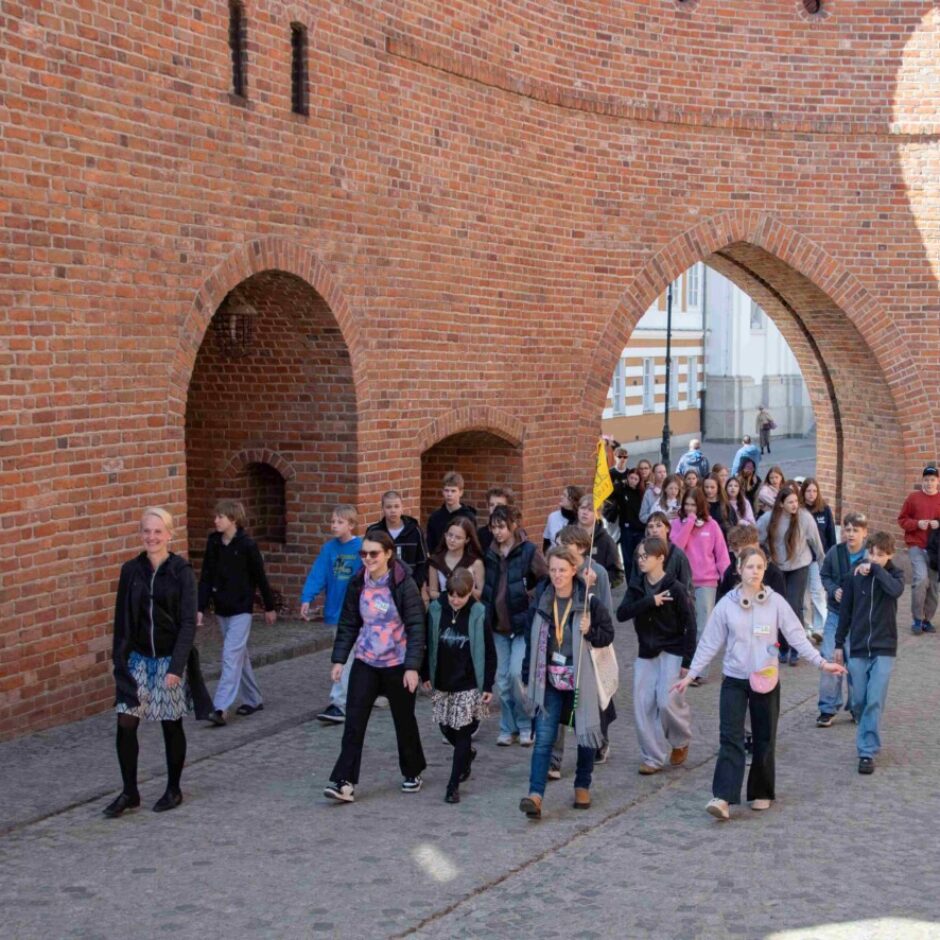 Liela skolēnu un pieaugušo grupa, ko vada pieaugušais ar dzeltenu karogu, staigā pa bruģētu ielu gar sarkanu ķieģeļu sienu ar arkām.