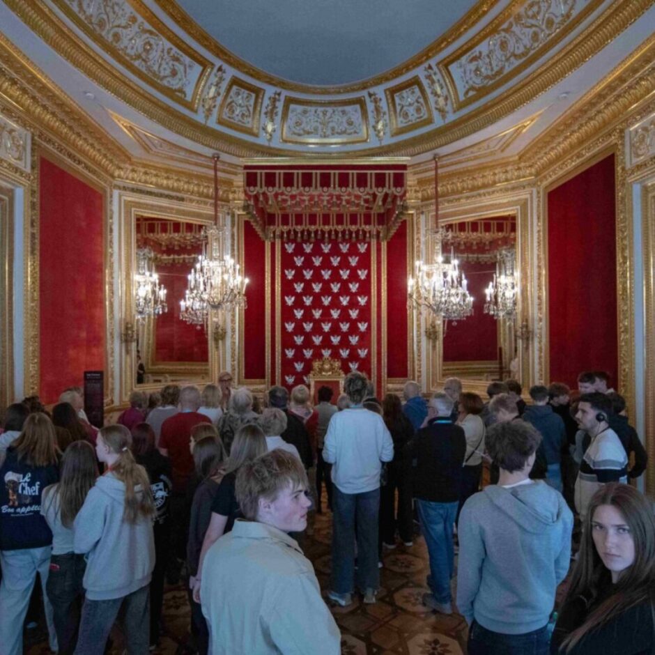 Liela cilvēku grupa atrodas greznā zālē ar sarkanām sienām, bagātīgiem zelta rotājumiem un augstiem, velvētiem griestiem. Karājas kristāla lustras, bet zāles galā ir izcils sarkans un zeltains ceremoniālais elements ar atkārtotiem baltiem simboliem. Daudzi grupas dalībnieki, daži ar austiņām, skatās uz šo elementu.