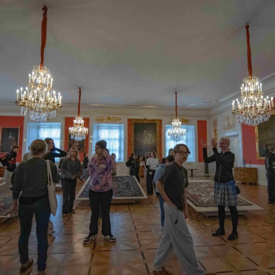 Telpā ar augstiem griestiem, sarkanām sienām un vairākām spīdošām kristāla lustrām grupa cilvēku apskata izliktus paklājus uz paaugstinājumiem. Sienas rotā gleznas zelta rāmjos un zeltaini ornamenti.