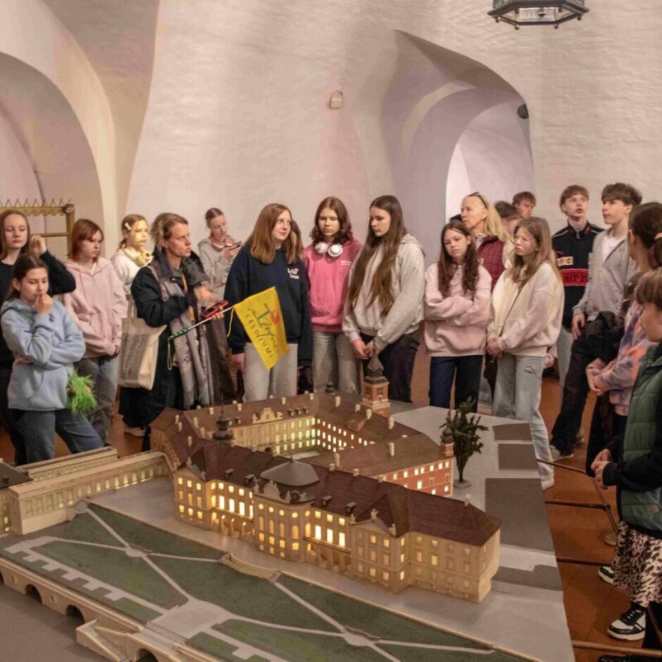 Gide vada jauniešu grupu pa telpu ar baltām velvētām sienām, kurā aplūkojams liels pils vai ēku kompleksa modelis ar apgaismotiem logiem un dārza plānojumu. Gide tur dzeltenu karodziņu ar sarkanu sirds simbolu.