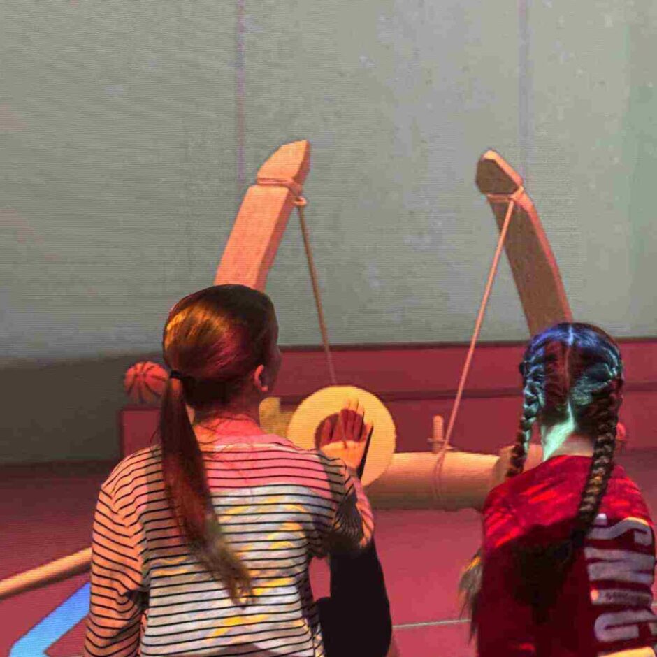 Divas meitenes skatās uz lielu, projicētu koka katapultas attēlu uz sienas. Meitene kreisajā pusē ir svītrainā kreklā, bet meitene labajā pusē — sarkanā kreklā ar vārdu 