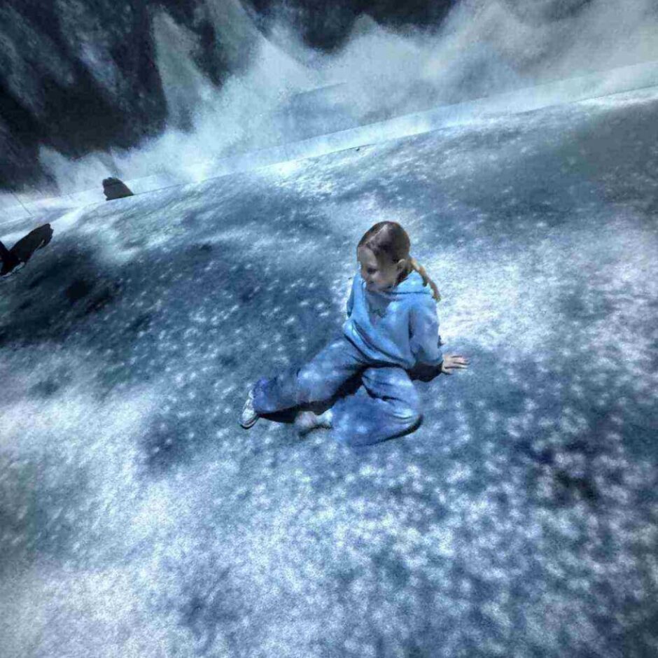 Jauna meitene gaišā zilā sporta tērpā sēž uz grīdas, ko pārklāj zilas un baltas ūdens un ledus efekta projekcijas, ar lieliem viļņiem uz sienas fonā.