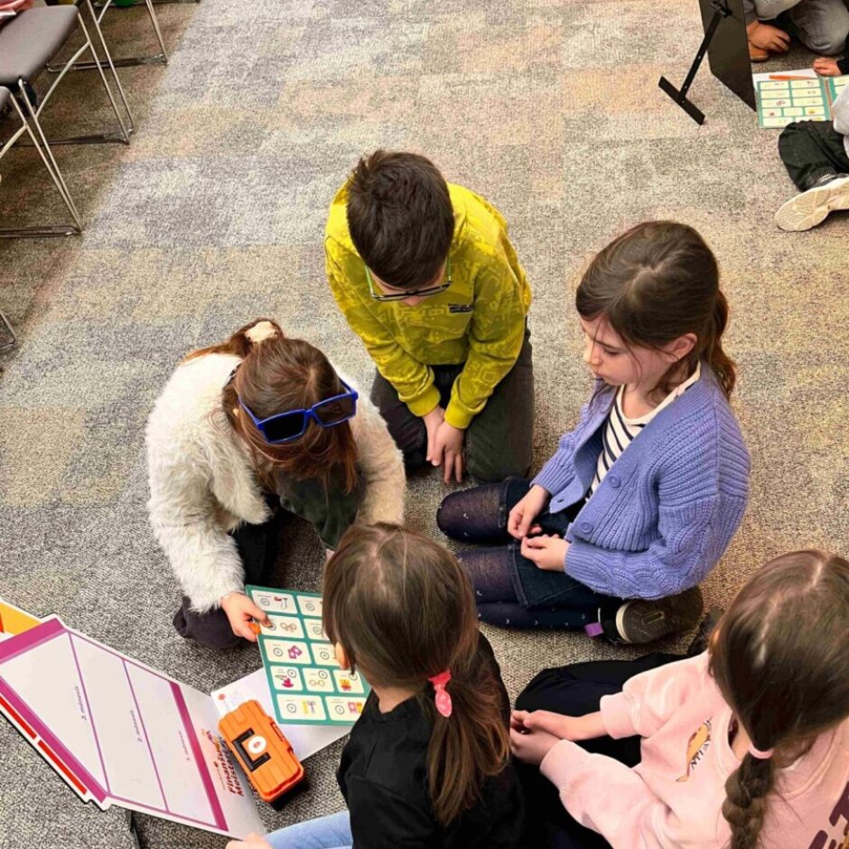 Četri bērni sēž uz grīdas, iesaistoties programmēšanas spēlē ar simbolu režģi, oranžu rotaļu robotu un atvērtu instrukciju dēli, uz kura redzams teksts 