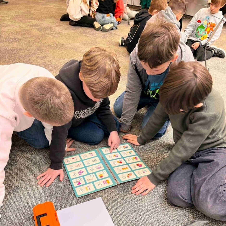 Četri zēni sēž uz pelēka paklāja un aktīvi darbojas ar atvērtu, zilu grāmatu, kurā redzami attēli un simboli. Viens no zēniem rāda ar pirkstu uz grāmatu. Blakus atrodas oranža kaste un balta papīra lapa. Fonā sēž citi bērni.