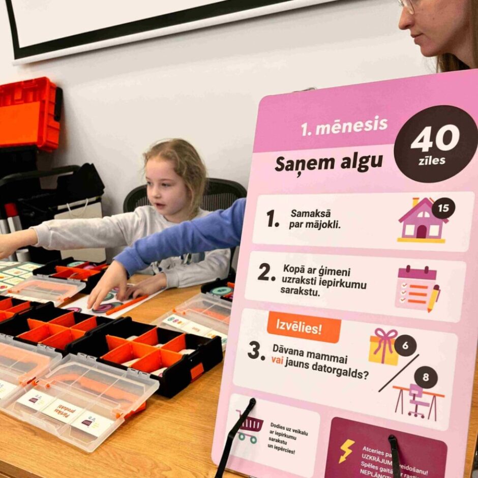 Jauna meitene ar blondiem matiem piedalās izglītojošā spēlē pie koka galda, sniedzoties pēc spēles elementiem no caurspīdīgām kastēm. Pie galda ir rozā stends ar uzrakstiem: 